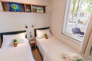 A hotel room with two beds, a shelf with hats, a window with a view, and a small table with a toy.