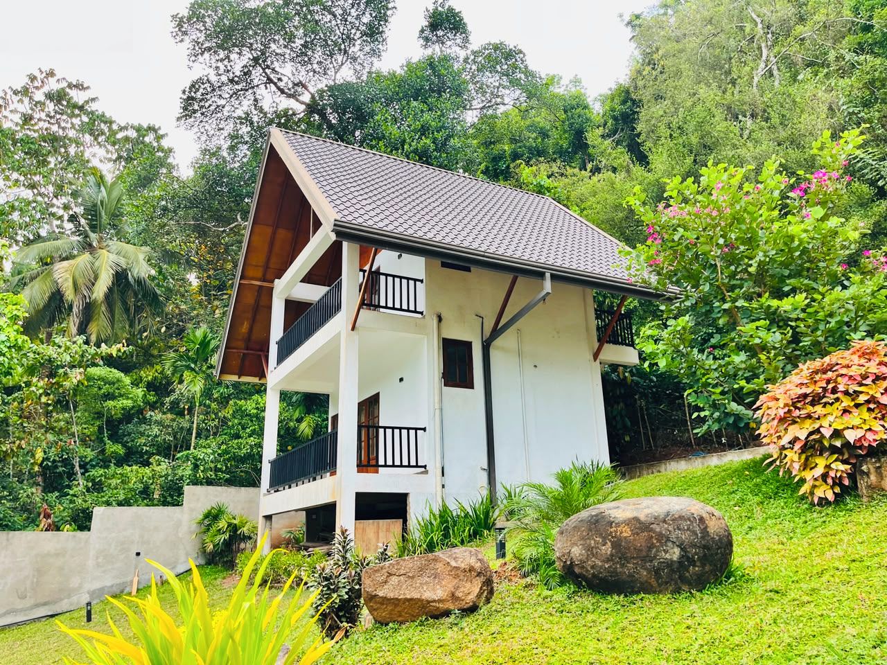 A two-story building with a balcony, surrounded by lush greenery and large rocks.