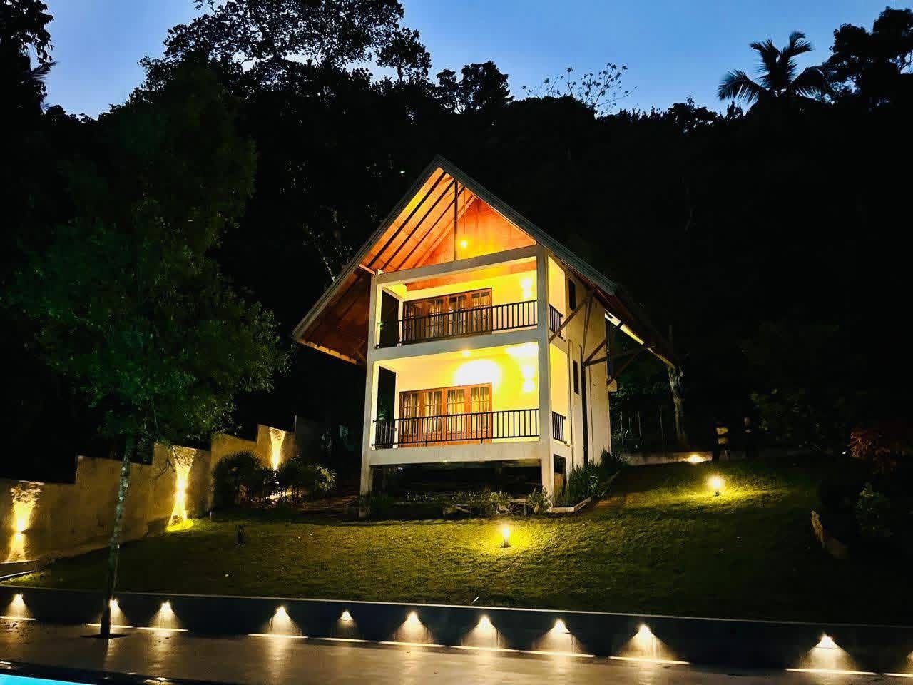 A two-story house with a balcony and a lit interior, surrounded by trees and a well-lit lawn.
