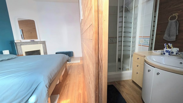 A bedroom with a bed, a bedside table, a nightstand, a wooden floor, a bathroom with a sink, a mirror, and a shower. - New Listing (Royan)