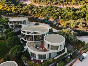 A modern, multi-level circular building with outdoor terraces, surrounded by lush greenery and a hilly landscape. - The Casa Cala Hotel (Bodrum)