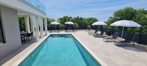 A rectangular swimming pool with clear water, surrounded by lounge chairs and umbrellas, and a patio area with a table and chairs. - Villa La Vie with heated pool (Garica)