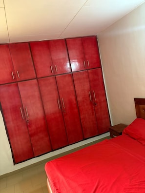 A room with a large red wardrobe, a bed with red bedding, and a small wooden nightstand. - New Listing (Abidjan)
