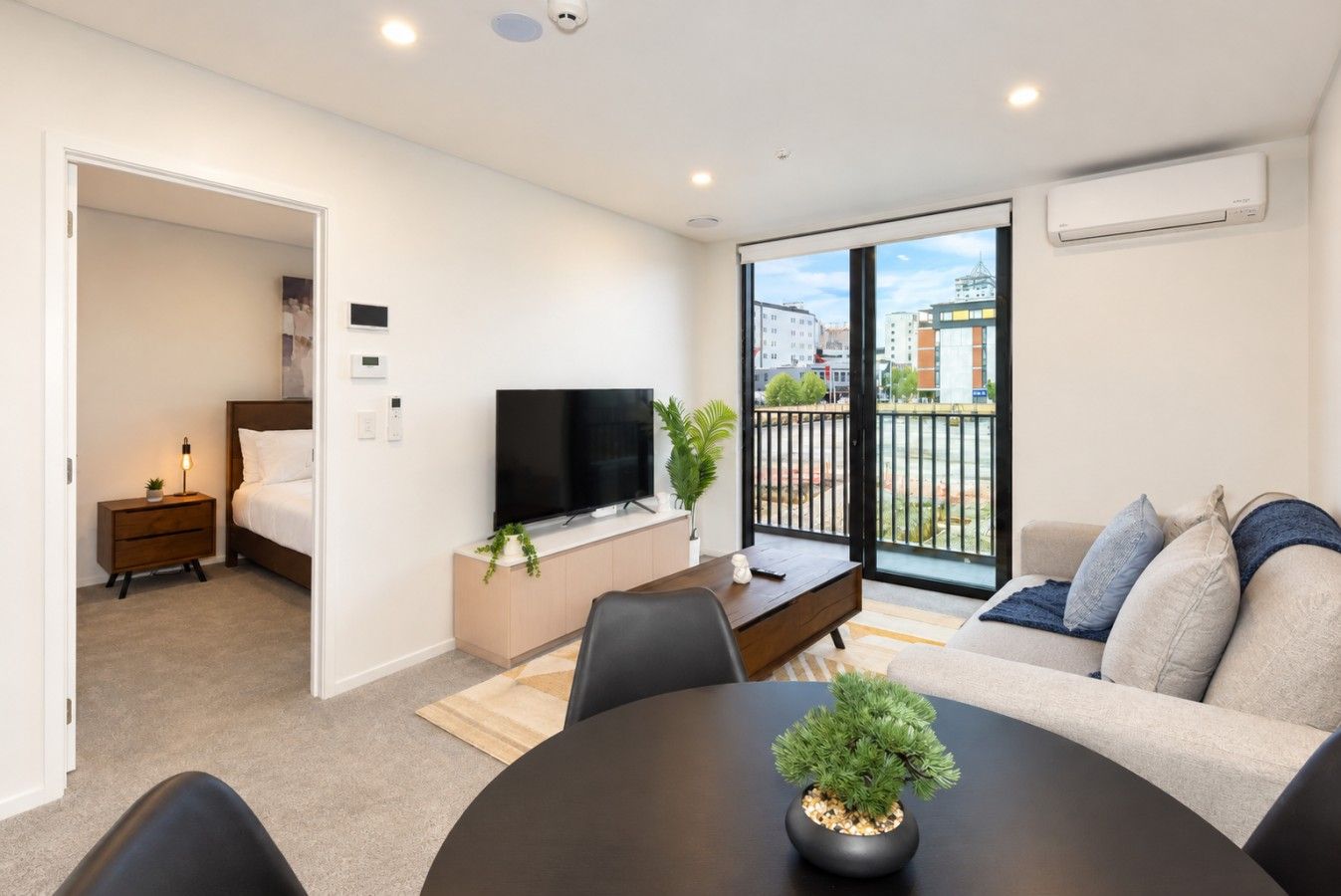 A modern living room with a sofa, a coffee table, and a TV. There's a balcony with a view of buildings.