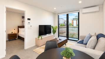 A modern living room with a sofa, a coffee table, and a TV. There's a balcony with a view of buildings.