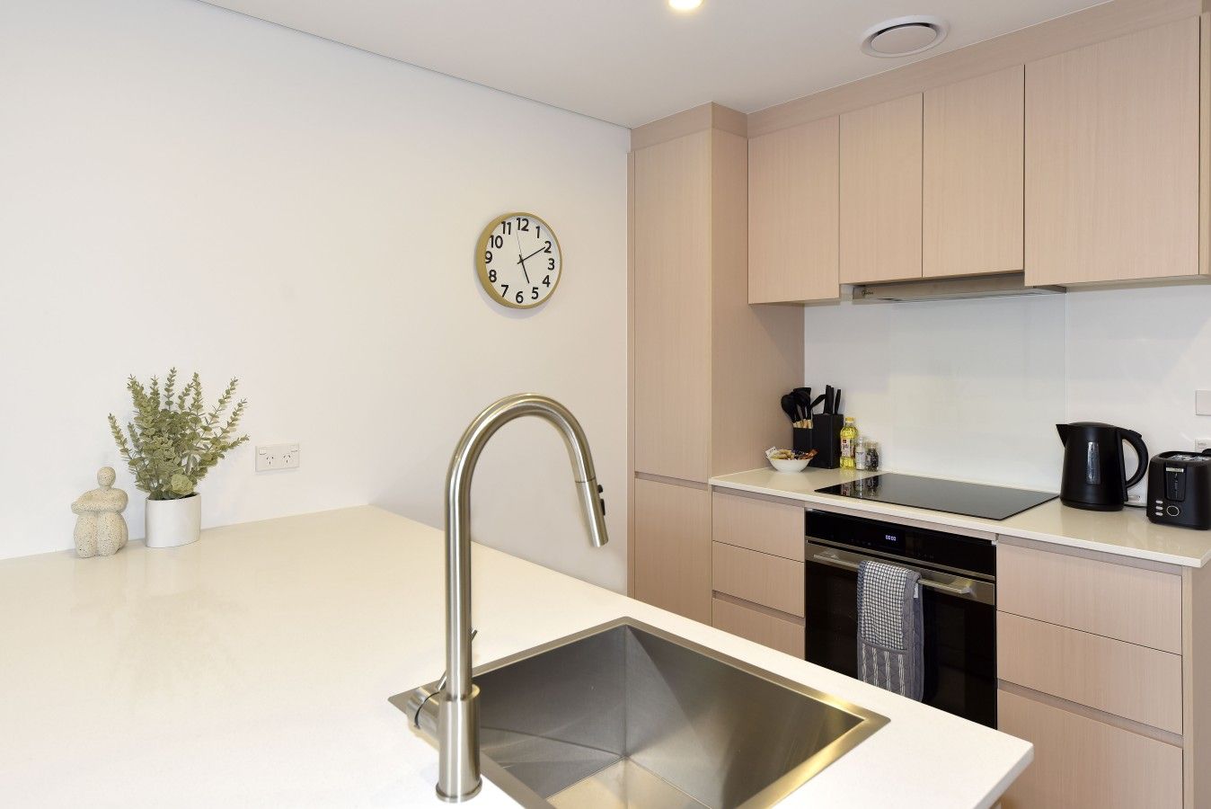 A modern kitchen with a stainless steel sink, a wall clock, and light-colored cabinets.