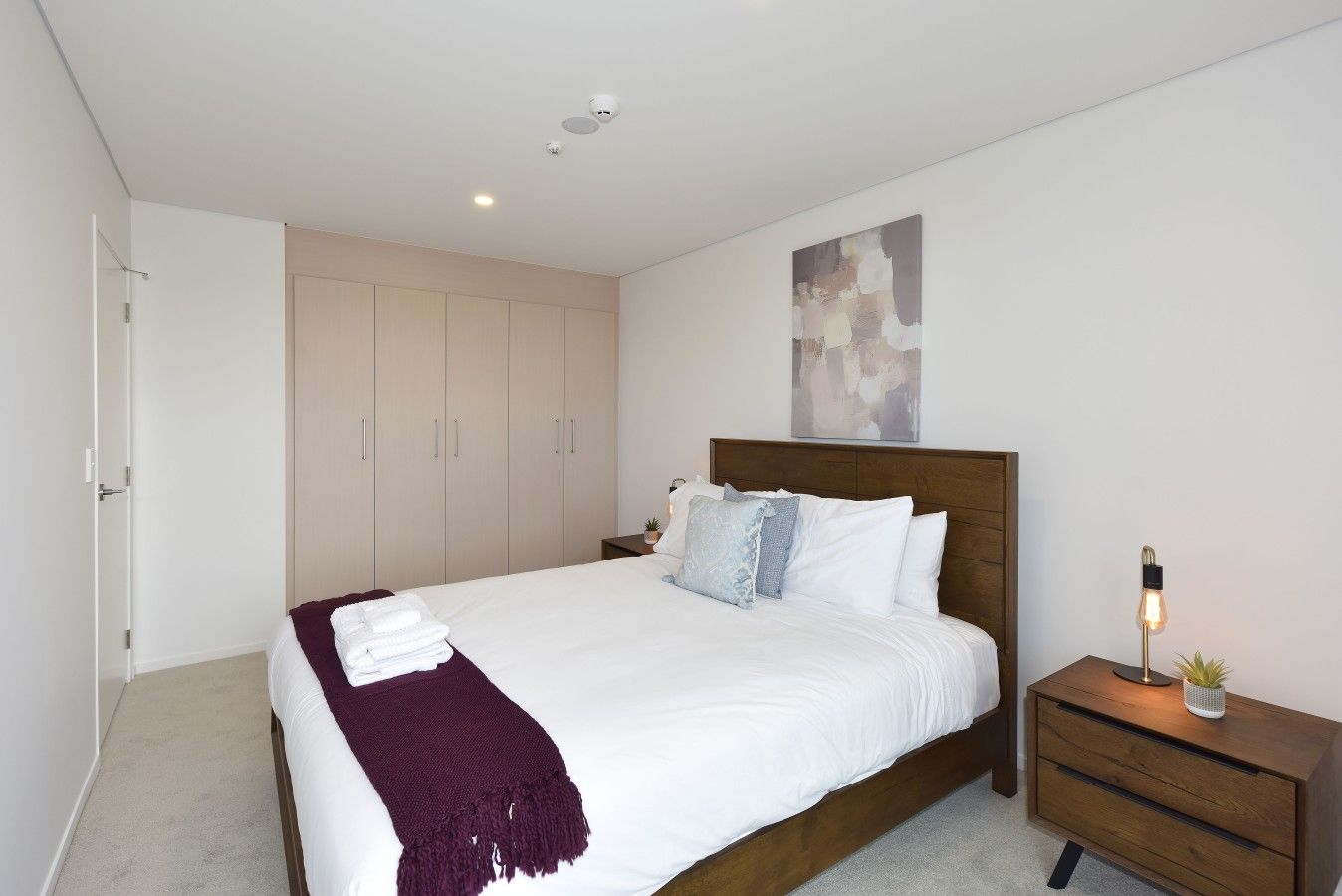 A modern bedroom with a large bed, a wooden headboard, a nightstand with a lamp, and a wardrobe.