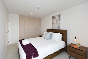 Une chambre à coucher moderne avec un grand lit, une tête de lit en bois, une table de chevet avec une lampe et une armoire.