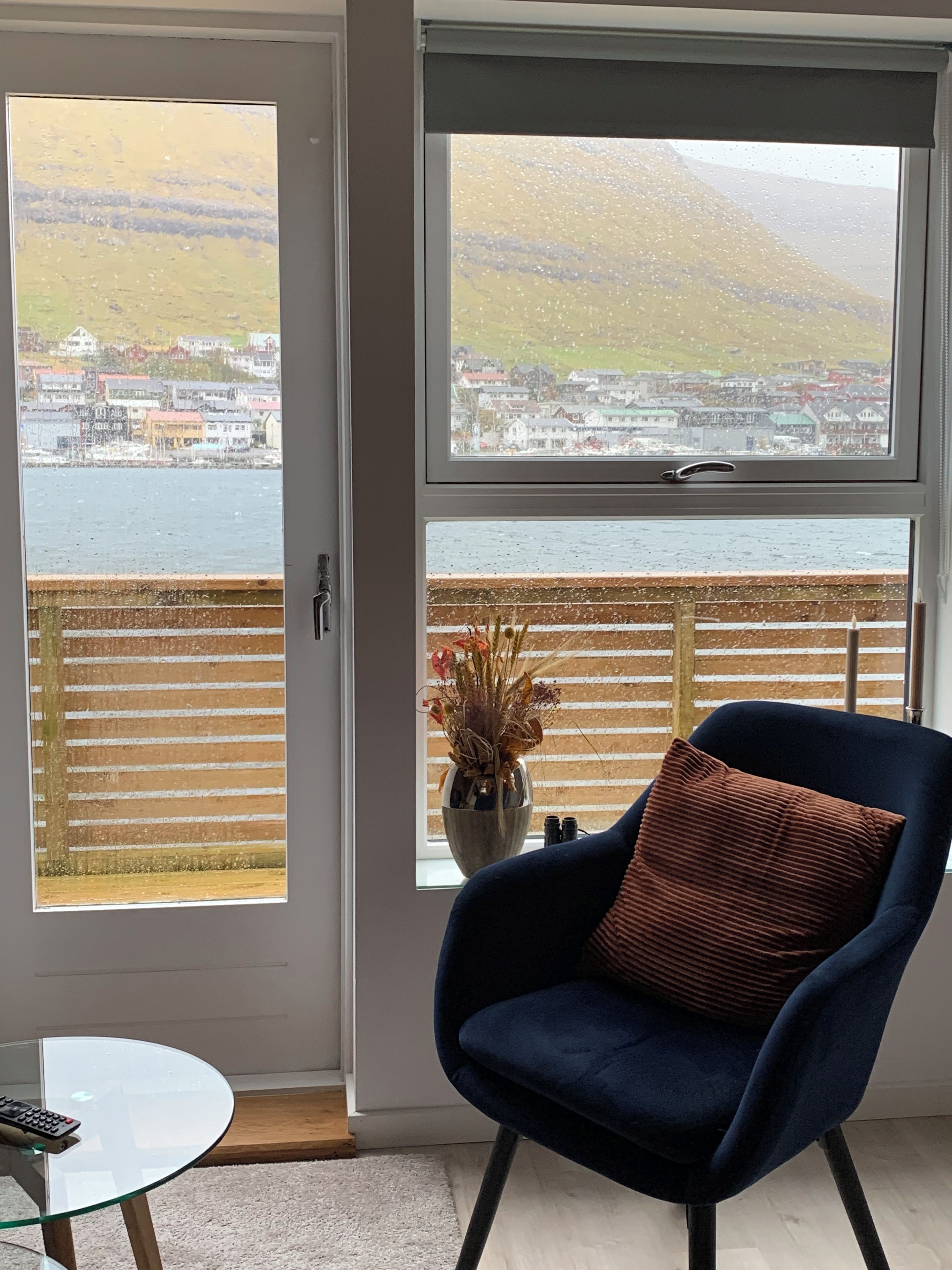 Una habitación con vista a un pueblo costero a través de un ventanal grande, una silla azul con cojín y una mesita con un control remoto.