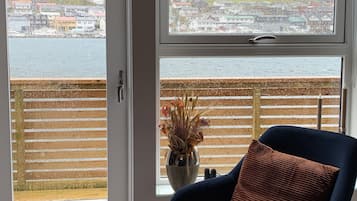 Una habitación con vista a un pueblo costero a través de un ventanal grande, una silla azul con cojín y una mesita con un control remoto.