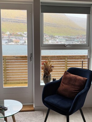 A room with a view of a coastal town through a large window, a blue chair with a cushion, and a small table with a remote control.