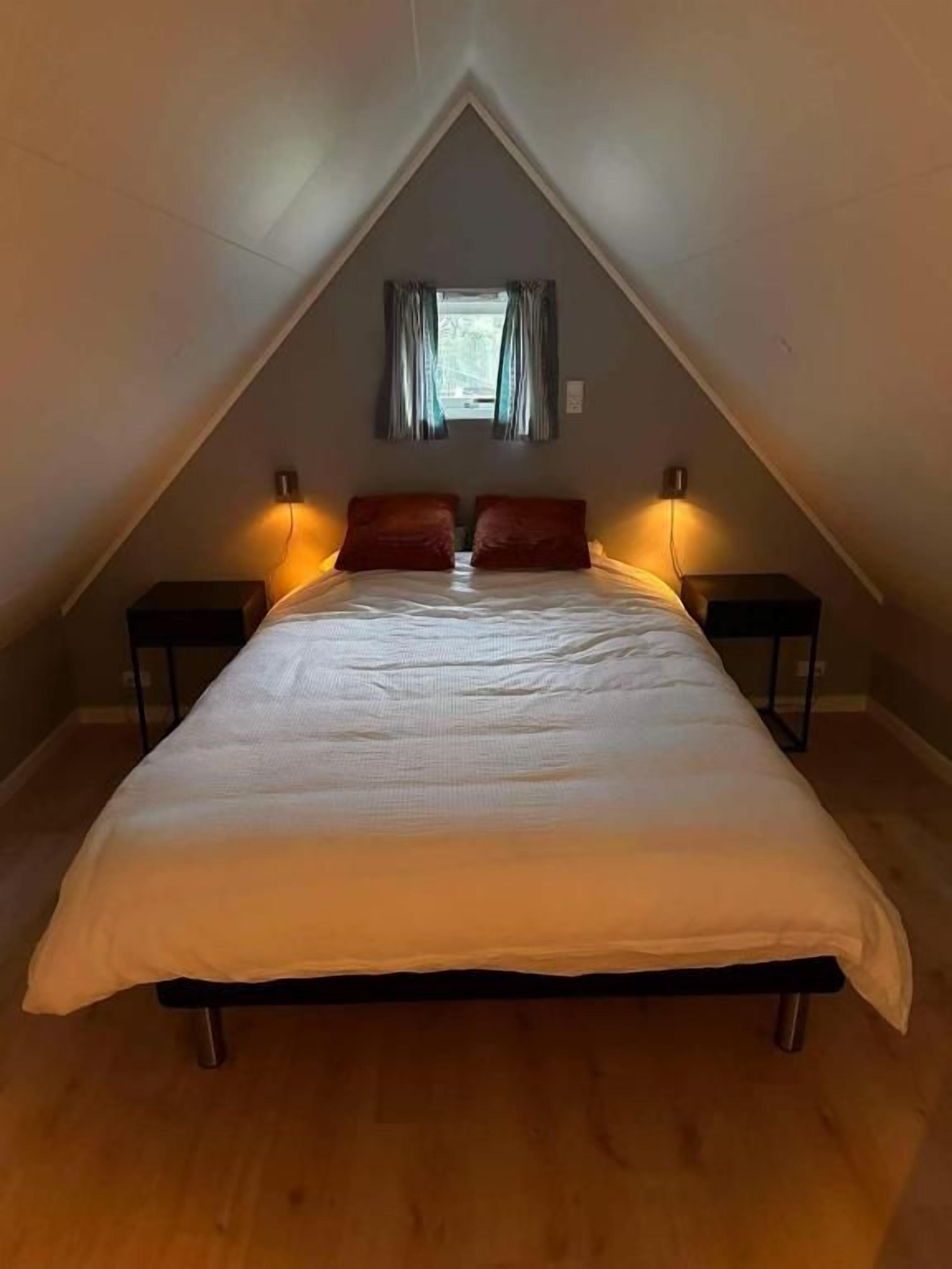 Un dormitorio con cama, dos mesitas de noche, una ventana con cortinas y techo inclinado.
