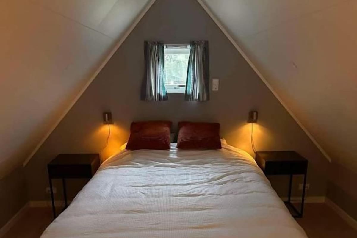 Un dormitorio con cama, dos mesitas de noche, una ventana con cortinas y techo inclinado.