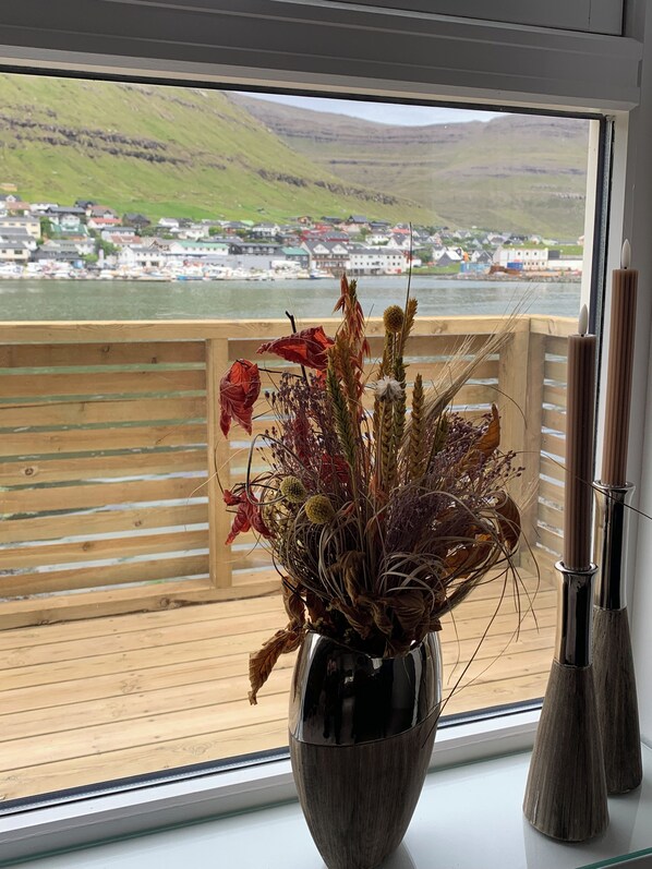 A window with a view of a coastal town, a wooden railing, and a vase with dried plants. - Klaksvíksvegur 64G (Klaksvík)