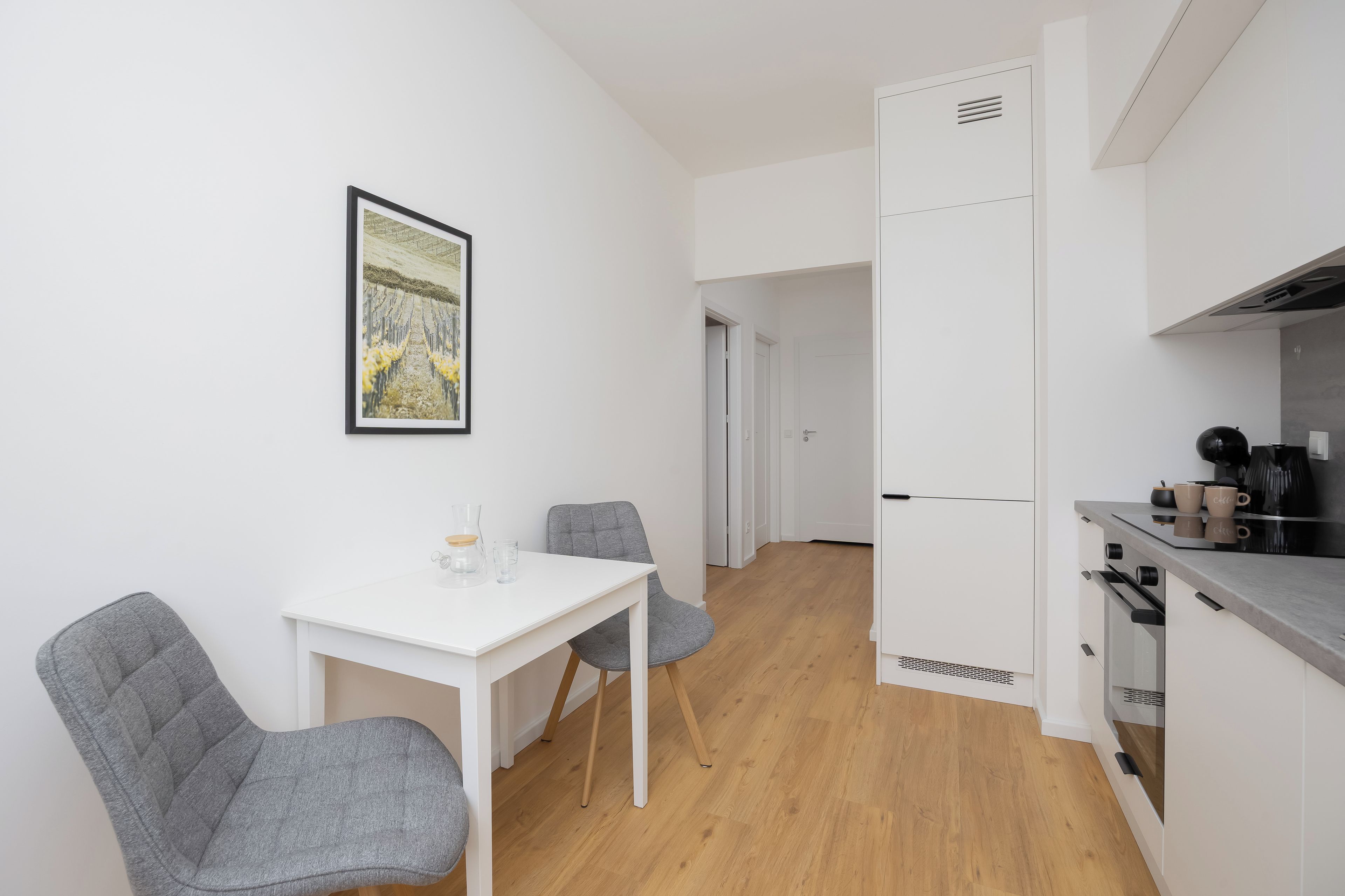 Cozinha compacta com mesa branca e duas cadeiras, um quadro na parede e vista para um quarto adjacente.