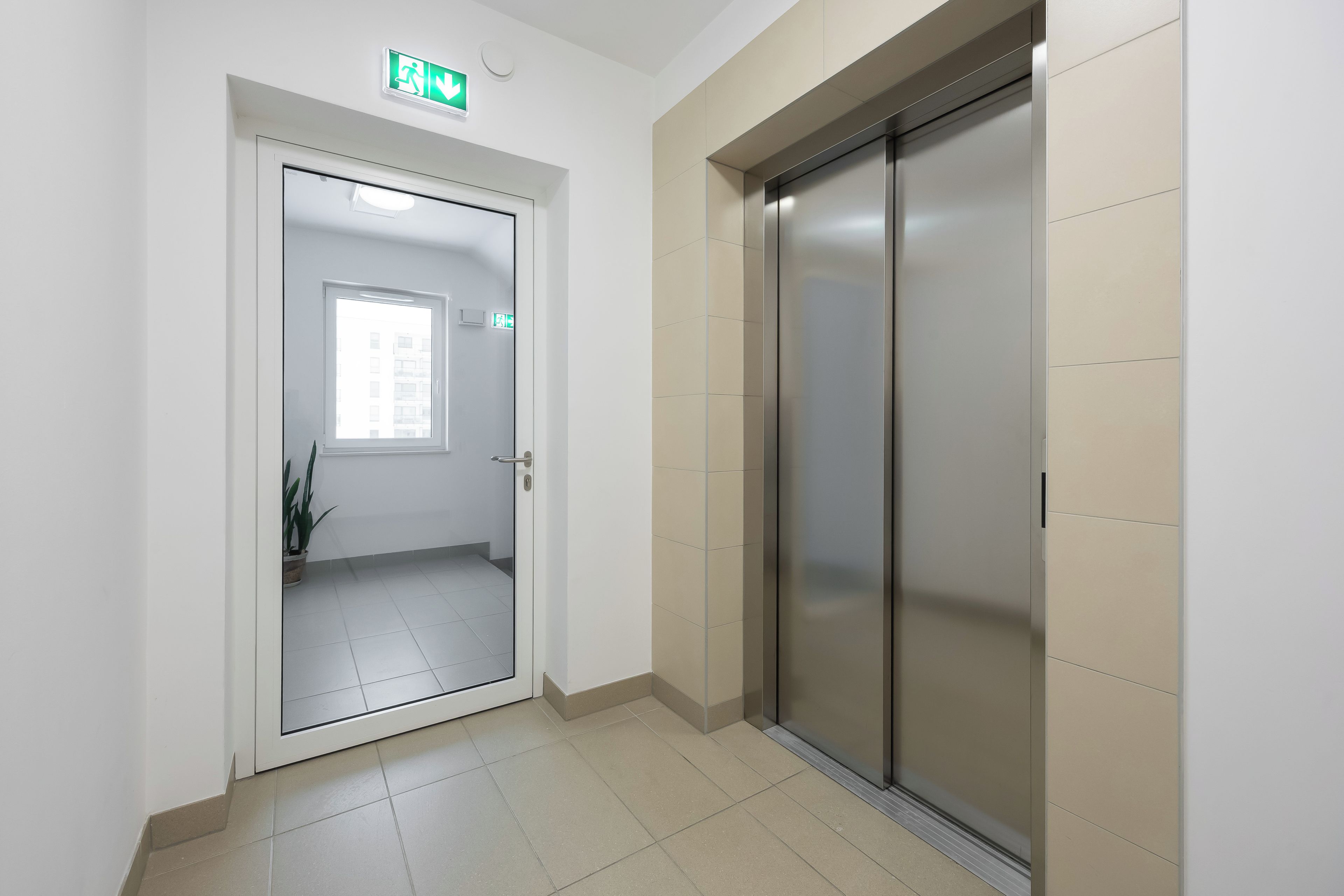 A hallway with a door leading to a room with a window, an emergency exit sign, and a wall-mounted elevator.