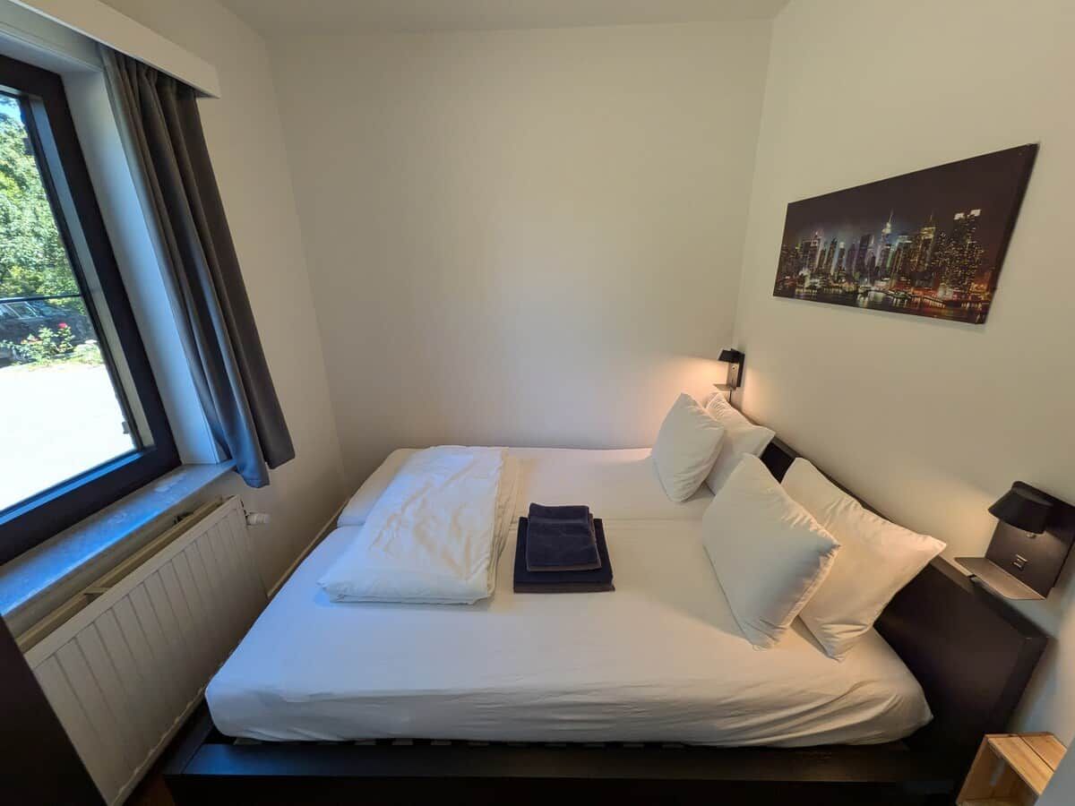Ein ordentlich bezogenes Bett mit Kissen und Decke, ein Nachttisch mit Lampe und ein Stadtbild an der Wand.