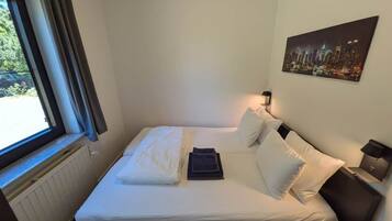 Ein ordentlich bezogenes Bett mit Kissen und Decke, ein Nachttisch mit Lampe und ein Stadtbild an der Wand.