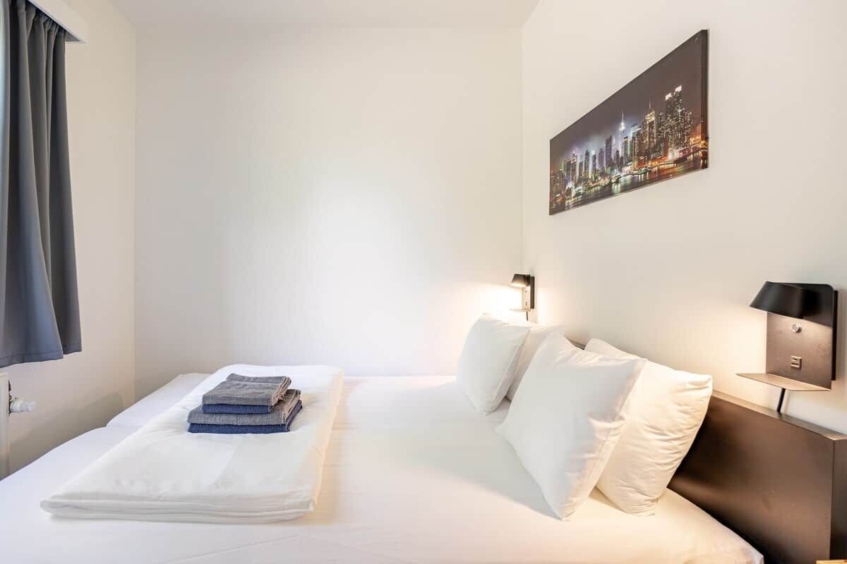 A hotel room with a bed, pillows, a nightstand, a lamp, and a cityscape painting on the wall.