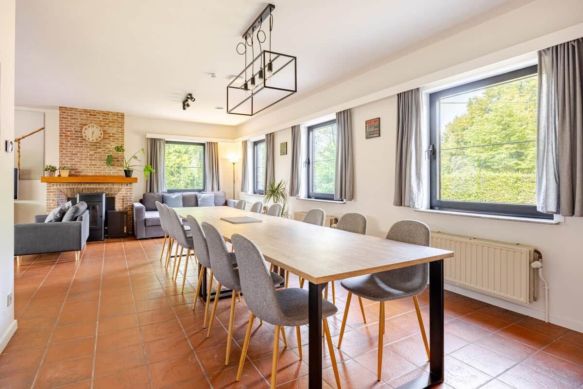 Un salon moderne avec une cheminée, un coin repas avec une longue table et des chaises, et de grandes fenêtres avec des rideaux.