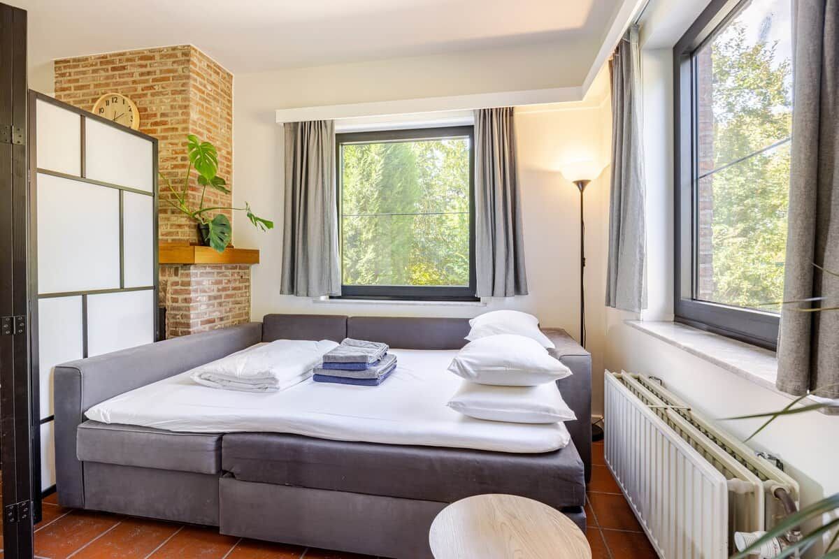 Een slaapkamer met een bed, een bakstenen muur, een raam met gordijnen, een radiator en een plant.