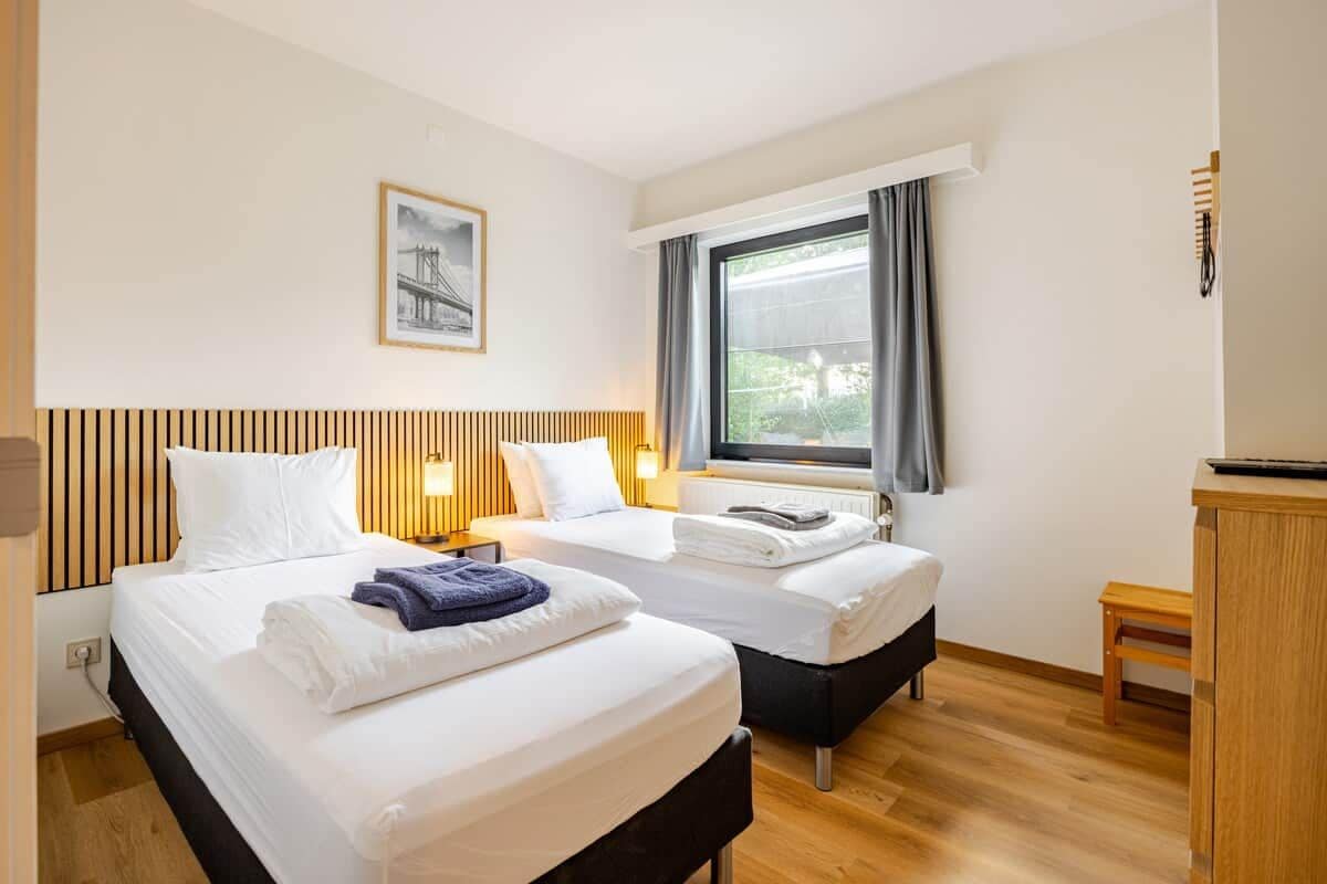 Une chambre d’hôtel avec deux lits, une fenêtre avec des rideaux, une tête de lit en bois, un tableau encadré au mur et une petite table en bois.
