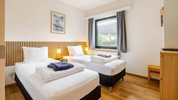 Une chambre d’hôtel avec deux lits, une fenêtre avec des rideaux, une tête de lit en bois, un tableau encadré au mur et une petite table en bois.