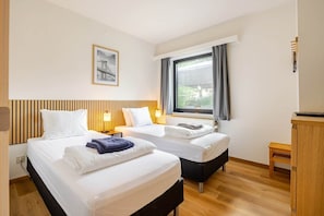 A hotel room with two beds, a window with curtains, a wooden headboard, a framed picture on the wall, and a small wooden table.
