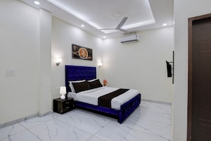 A bedroom with a bed, nightstand, wall-mounted lamp, ceiling fan, and an air conditioning unit. - Super Hotel O Clock Tower Dehradun (Dehradun)