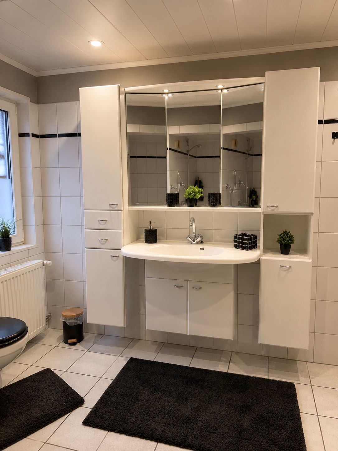 A modern bathroom with a white sink, mirror, and cabinets. There are black rugs on the floor and a small trash bin.