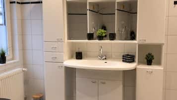 A modern bathroom with a white sink, mirror, and cabinets. There are black rugs on the floor and a small trash bin.