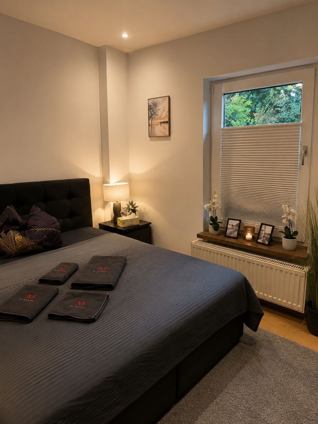 Et soveværelse med seng, sengeborde, et vindue med persienner, en radiator og en lille hylde med pynteting.