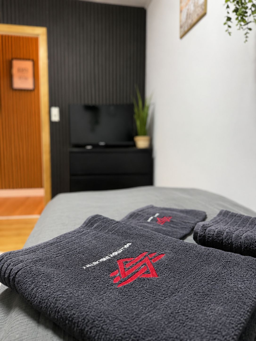 Una cama bien hecha con sábanas grises y un logo rojo, un mueble de televisión negro con una planta, y una puerta de madera en una habitación moderna.