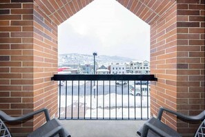 A balcony with brick walls and a metal railing, overlooking a street with buildings and a snow-covered landscape. - 2BR Winter Wonderland (Park City)