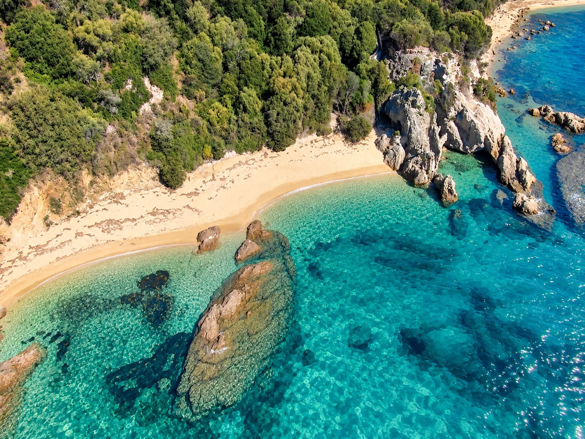 Luchtbeeld van een kustgebied met helder turquoise water, een zandstrand, rotsformaties en weelderige groene begroeiing.