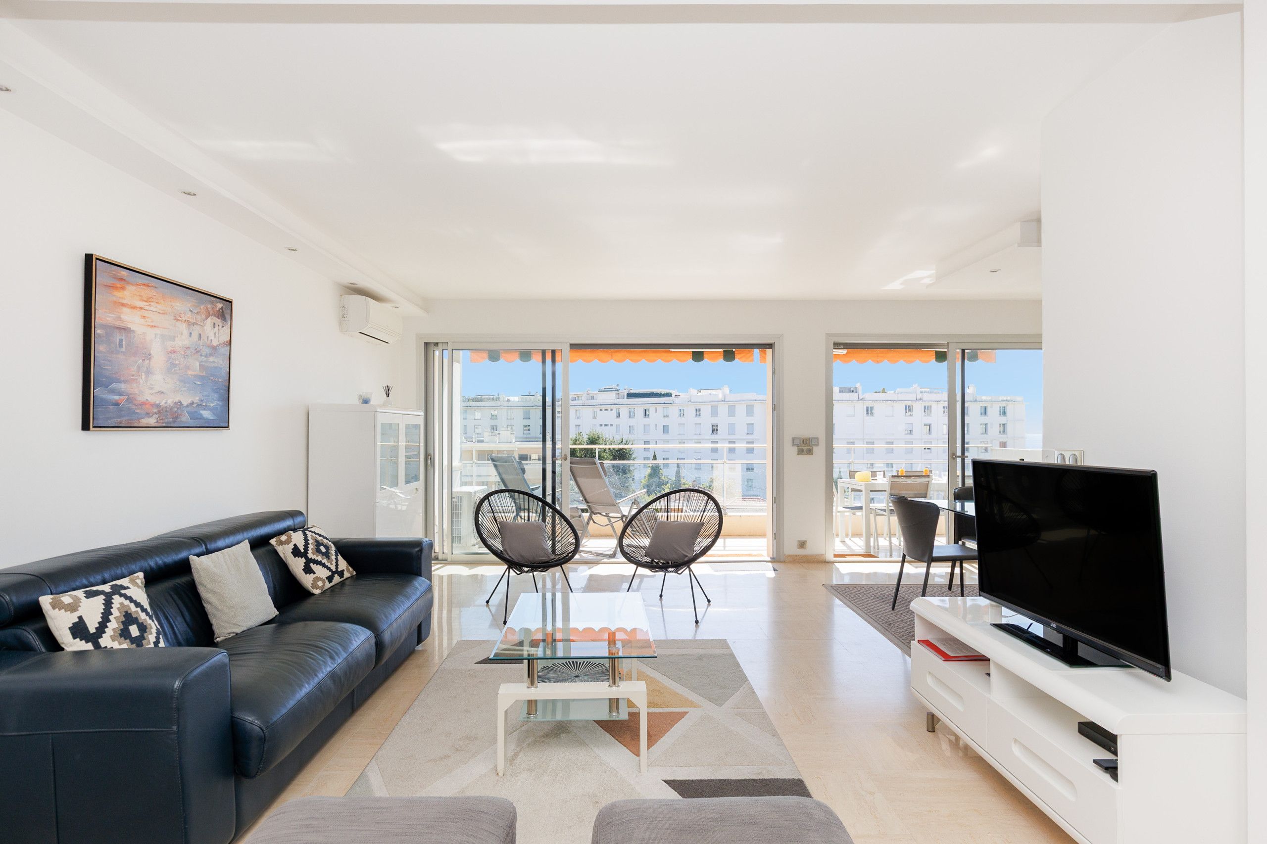A modern living room with a black leather sofa, a glass coffee table, two black chairs, a flat-screen TV, and a balcony with orange curtains.
