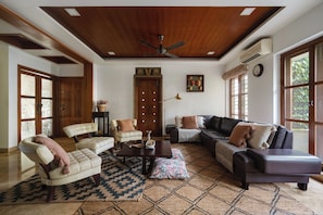 A spacious living room with a high ceiling, a ceiling fan, a large window, a sofa set, a coffee table, and a patterned rug.
