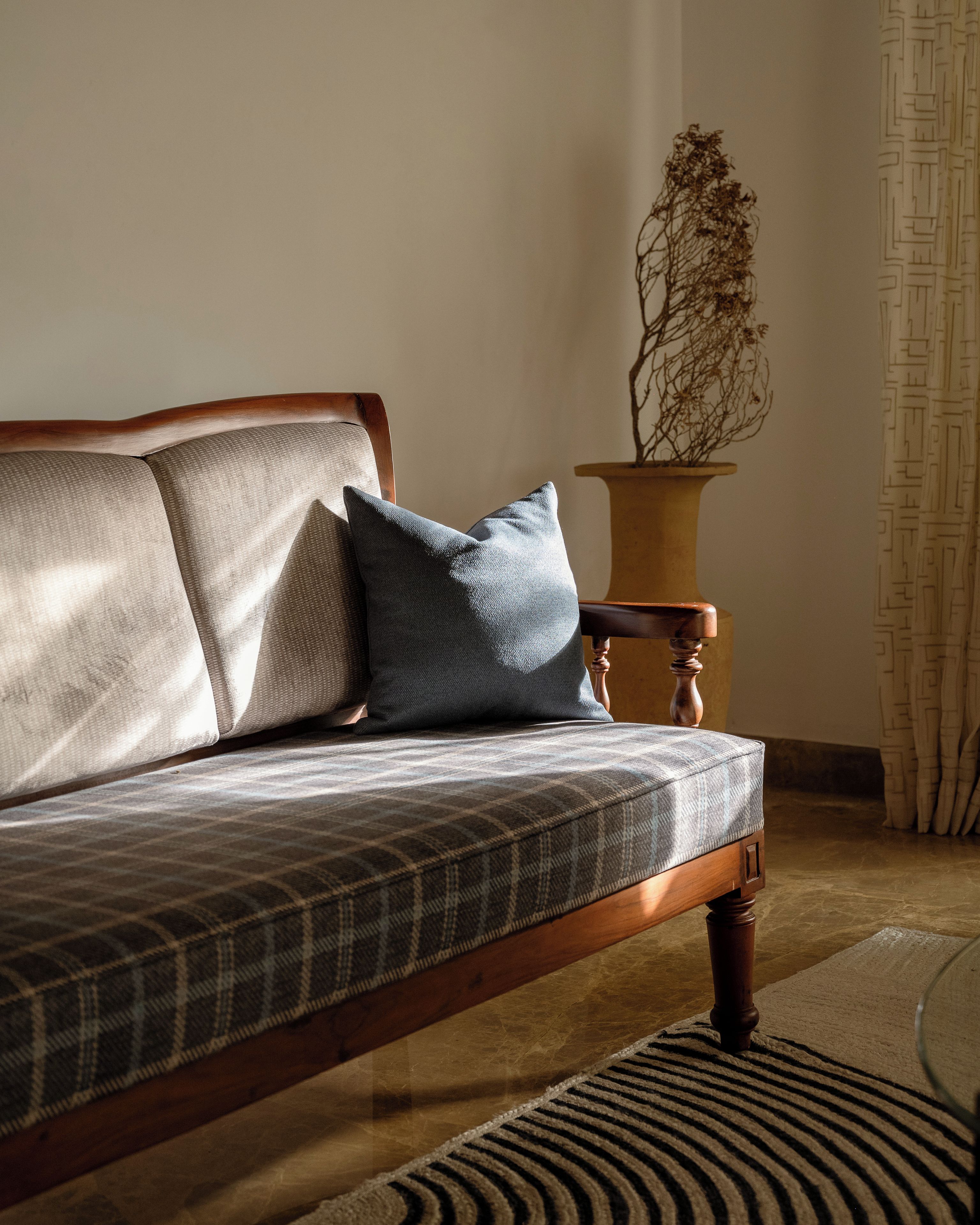 A wooden sofa with a plaid cushion, a dark blue pillow, and a potted plant with dried branches.