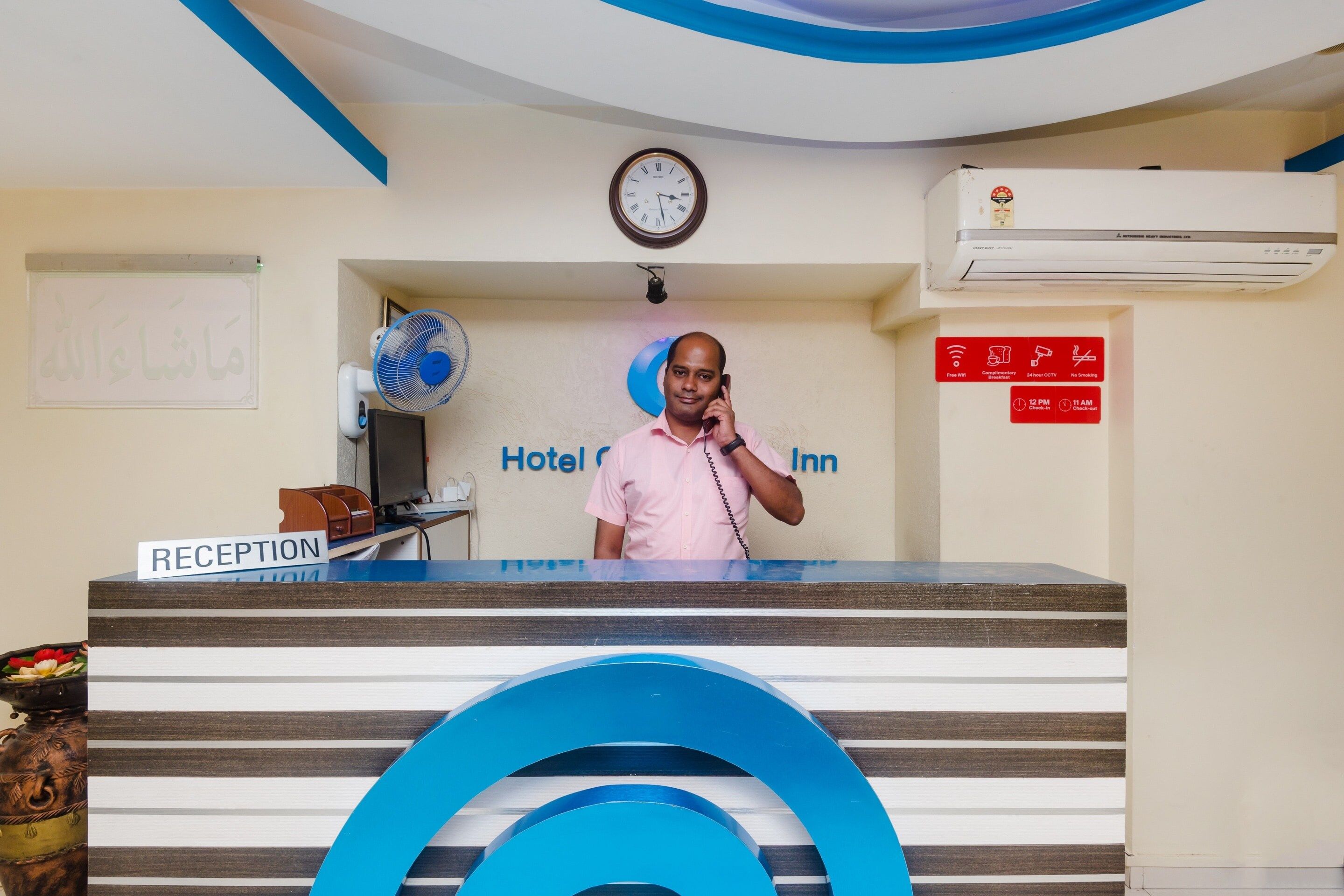 Un comptoir de réception d’hôtel avec une horloge, un panneau indiquant les heures d’arrivée et de départ, et un espace de réception équipé d’un ventilateur et d’un ordinateur.