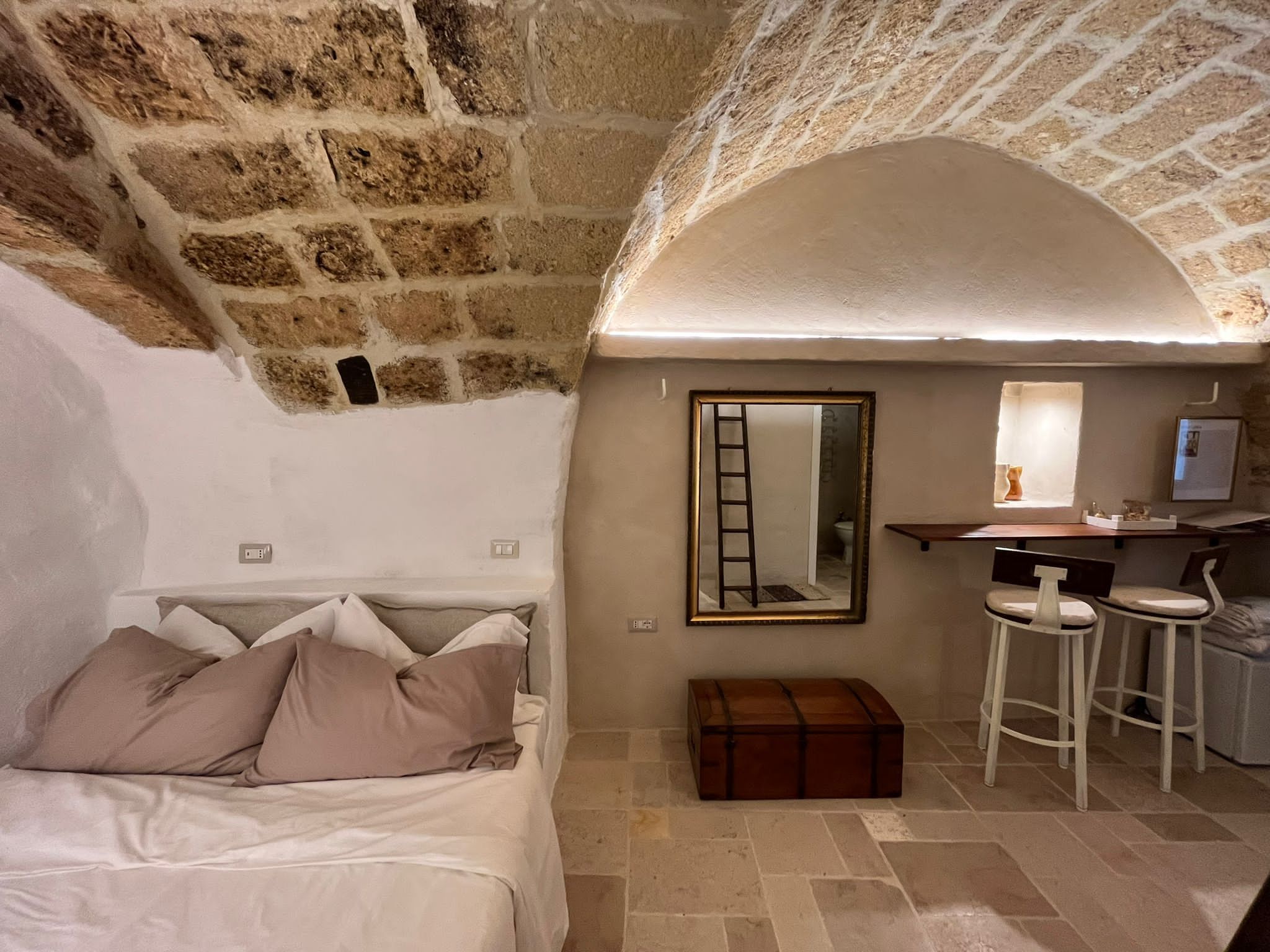 A cozy room with a stone wall, a bed with pillows, a small desk with a mirror, and a stool.