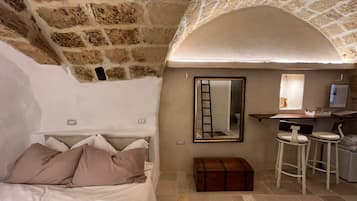 A cozy room with a stone wall, a bed with pillows, a small desk with a mirror, and a stool.