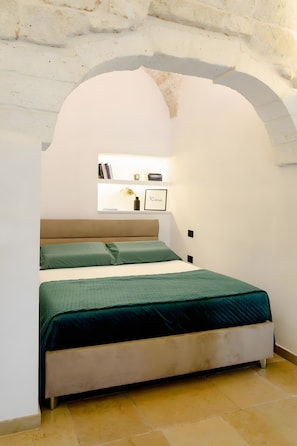 Una camera da letto con un letto, un comodino, uno scaffale con libri e un vaso, e una porta ad arco.