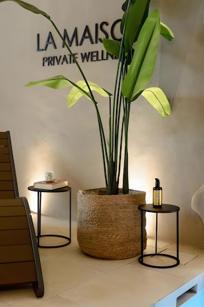 Un'area benessere privata con un cartello che riporta 'LA MAISON PRIVATE WELLNESS', una grande pianta in vaso e due tavolini con oggetti sopra.