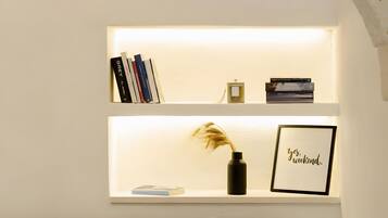 A bedroom with a bed, a green pillow, a shelf with books, a vase with dried plants, and a framed picture with text.