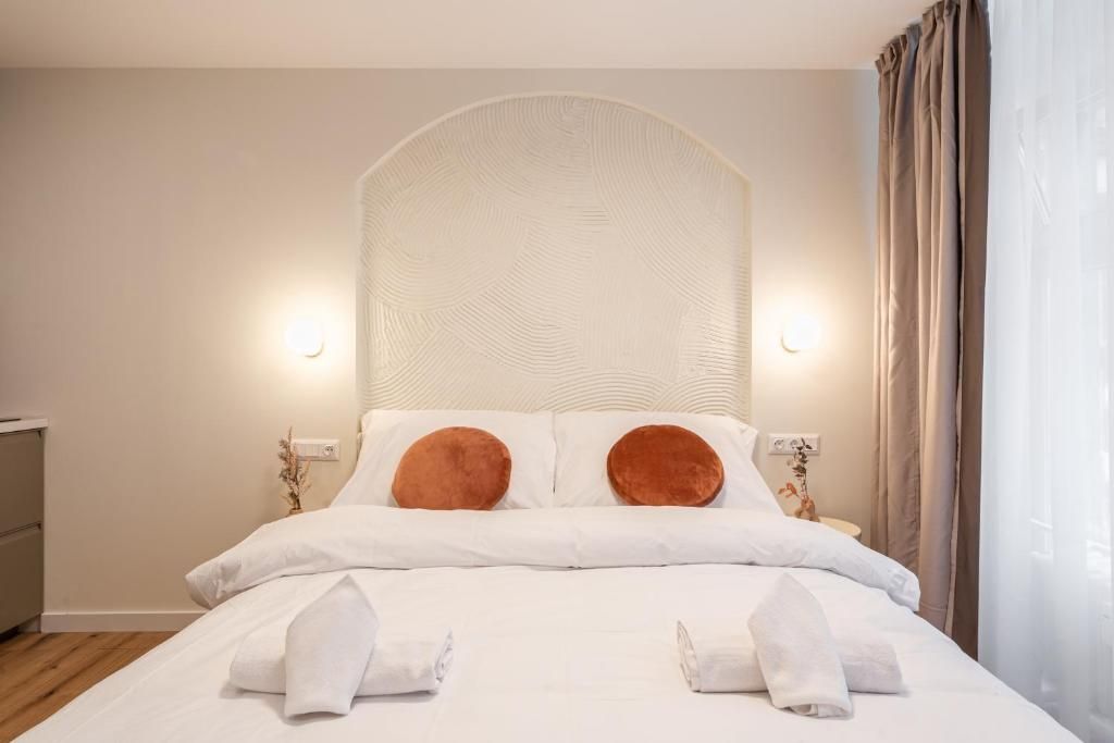 A neatly made bed with white linens and two brown pillows, a bedside table with a lamp, and a window with curtains.