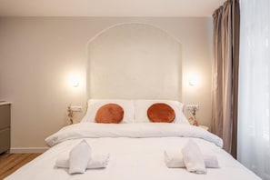 A neatly made bed with white linens and two brown pillows, a bedside table with a lamp, and a window with curtains. - Asbóth Boutique Suites by BQA (Budapest)