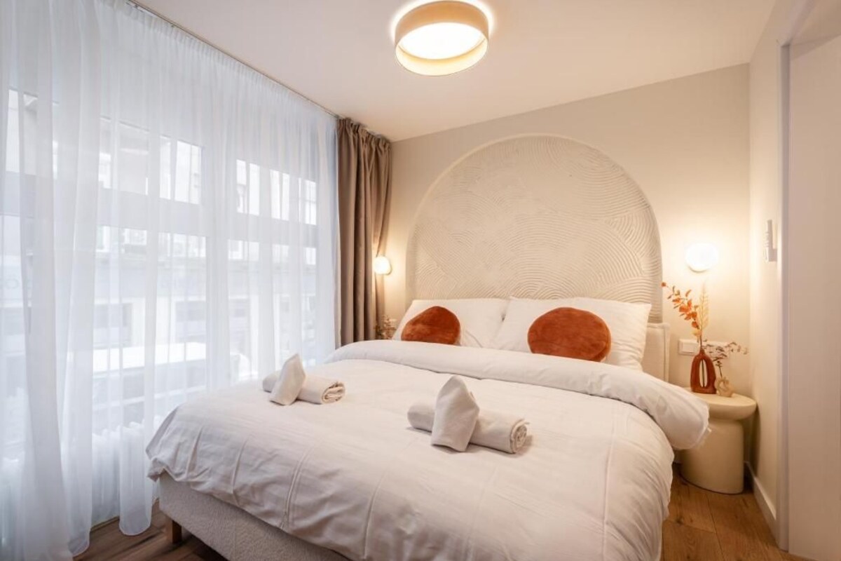A hotel room with a large bed, a round wall-mounted light fixture, and a bedside table with a vase.