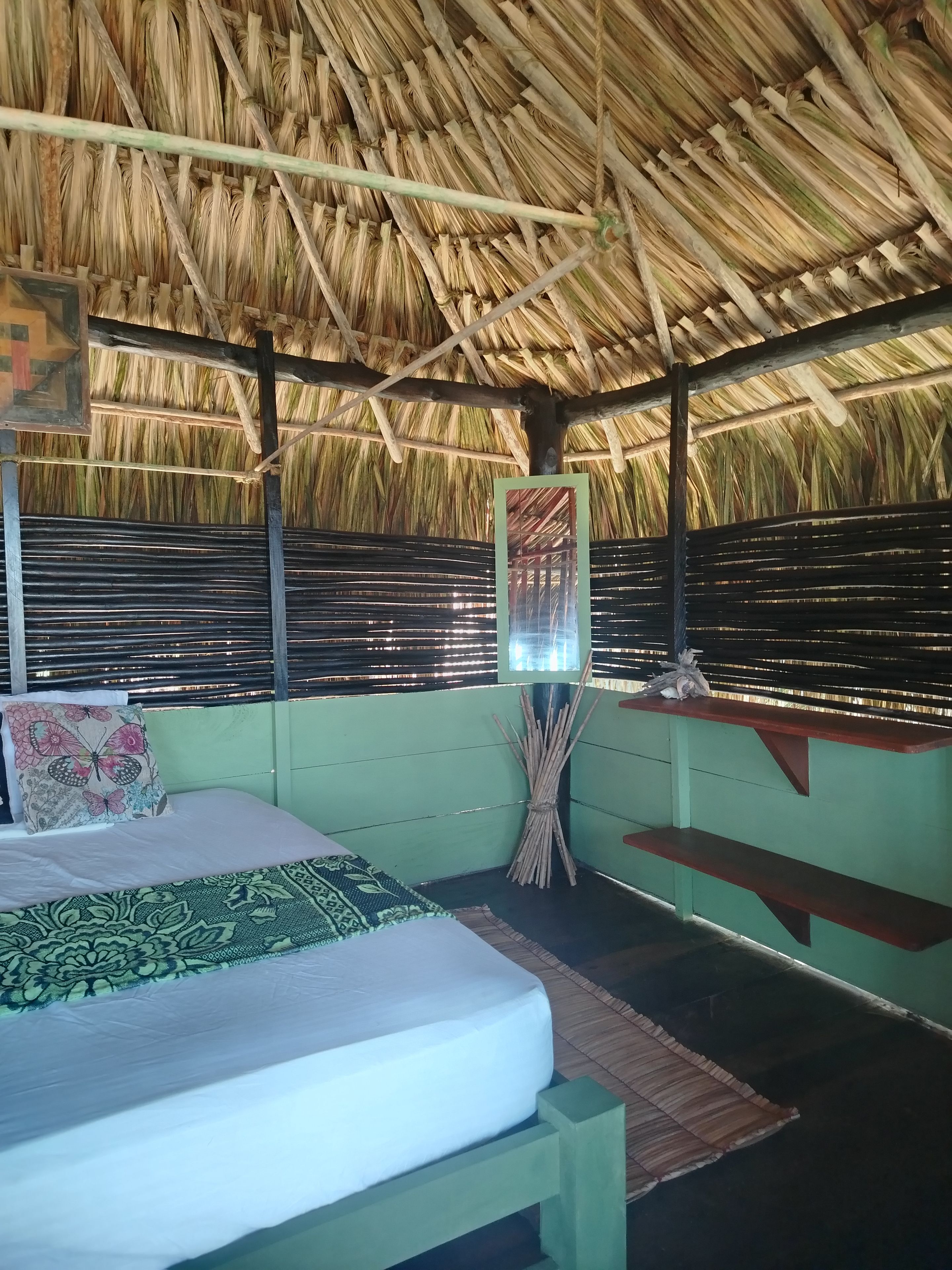 Una habitación con una cama, un banco, una cesta tejida y un techo de paja.