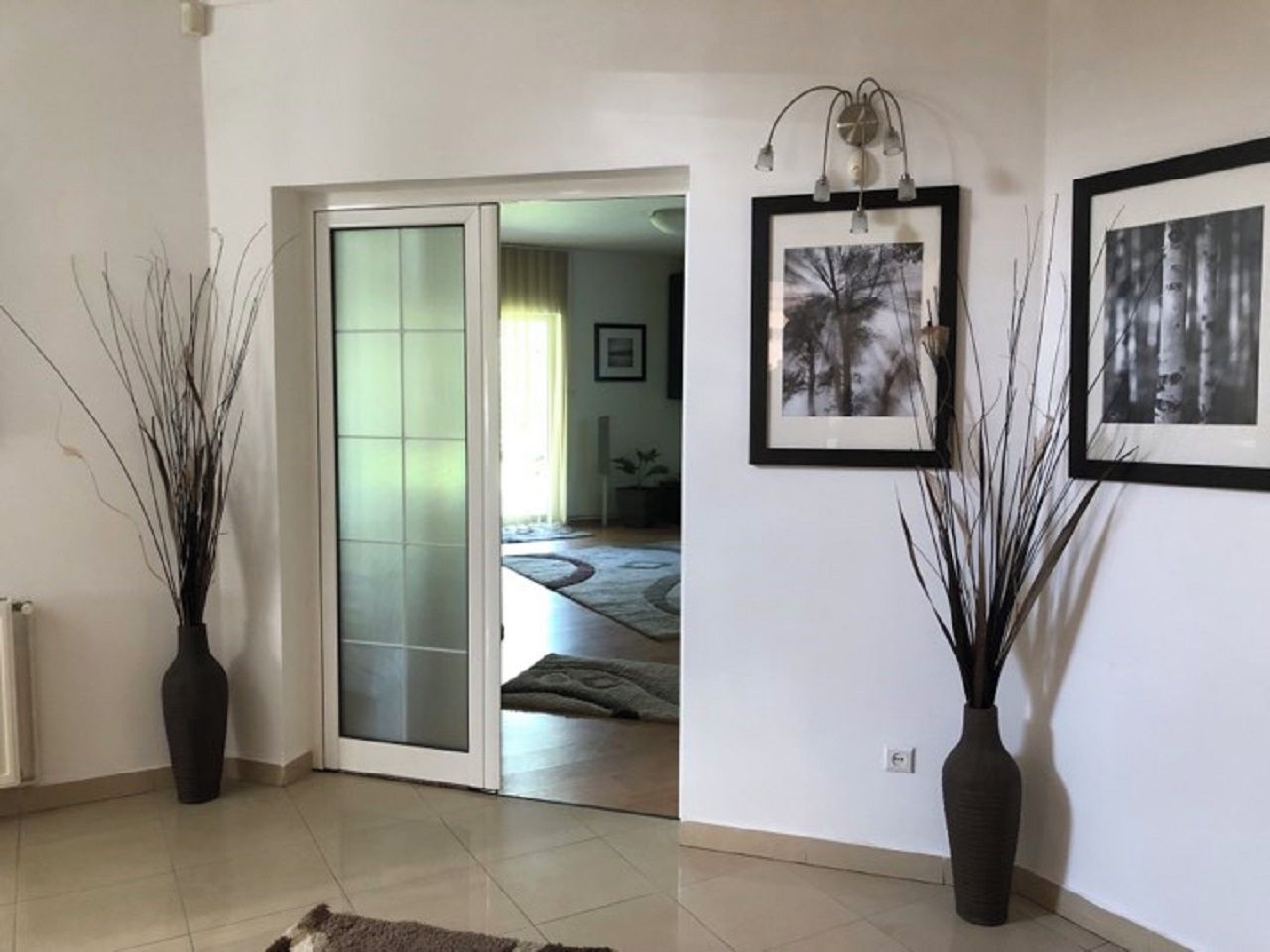 Un espacio interior con un divisor de vidrio corredizo que da a otra habitación, dos jarrones en el suelo y cuadros en la pared.