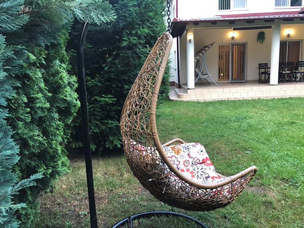 A hanging chair with a floral cushion in a grassy outdoor area with trees and a building in the background.